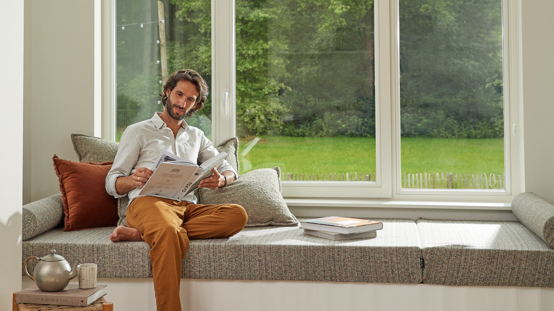 Hombre leyendo junto a un gran ventanal en una acogedora vivienda, que ilustra el confort y la eficiencia energética que proporcionan los equipos de aerotermia para reducir el impacto medioambiental en las soluciones de calefacción de interiores
