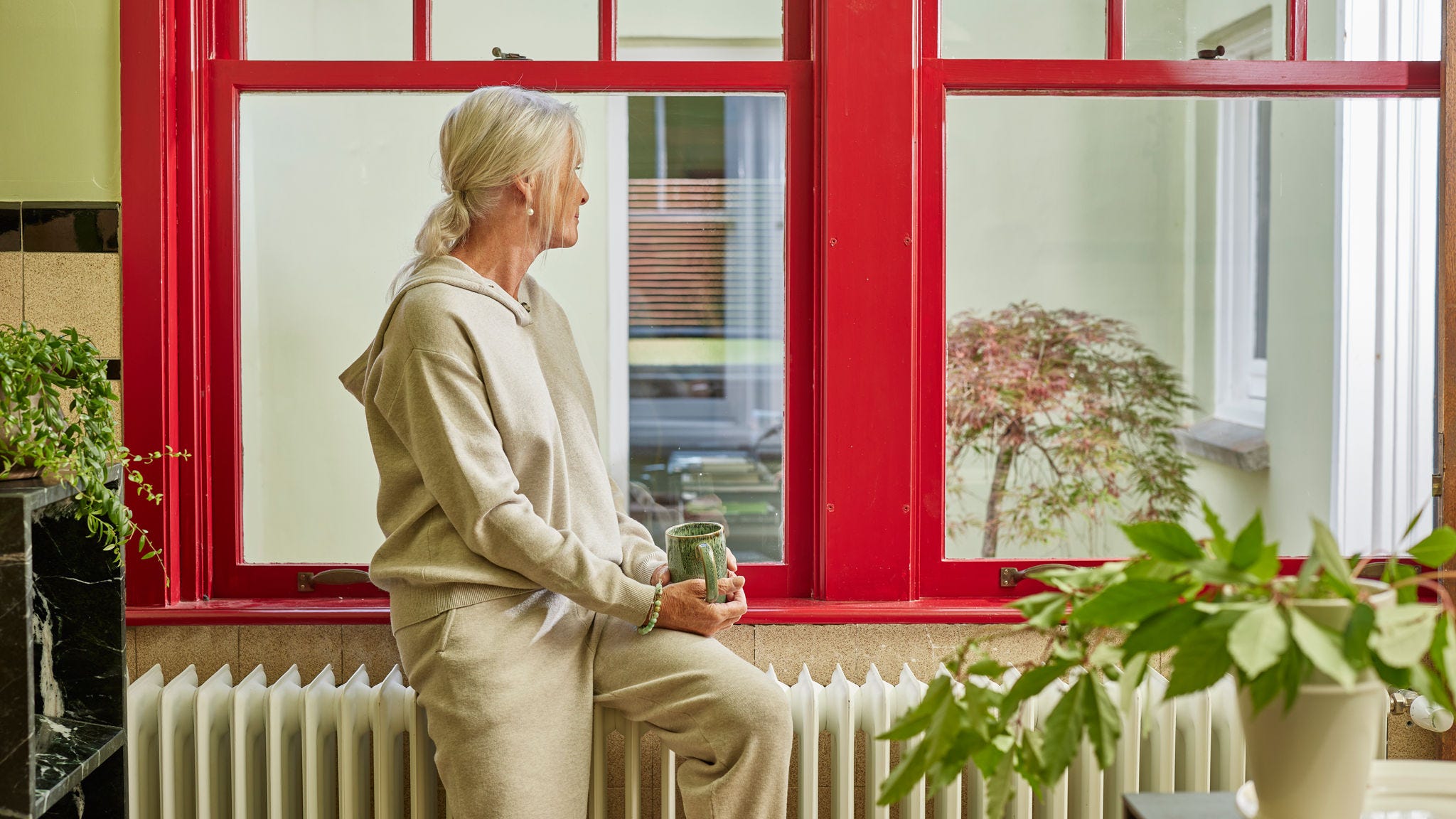 Mujer sentada en un radiador junto a un gran ventanal, disfrutando de un hogar cálido y energéticamente eficiente
