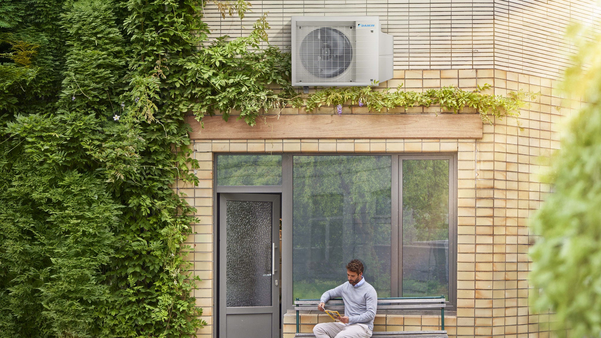 Un hombre leyendo en un banco frente a su casa, bajo un equipo de aerotermia Daikin.