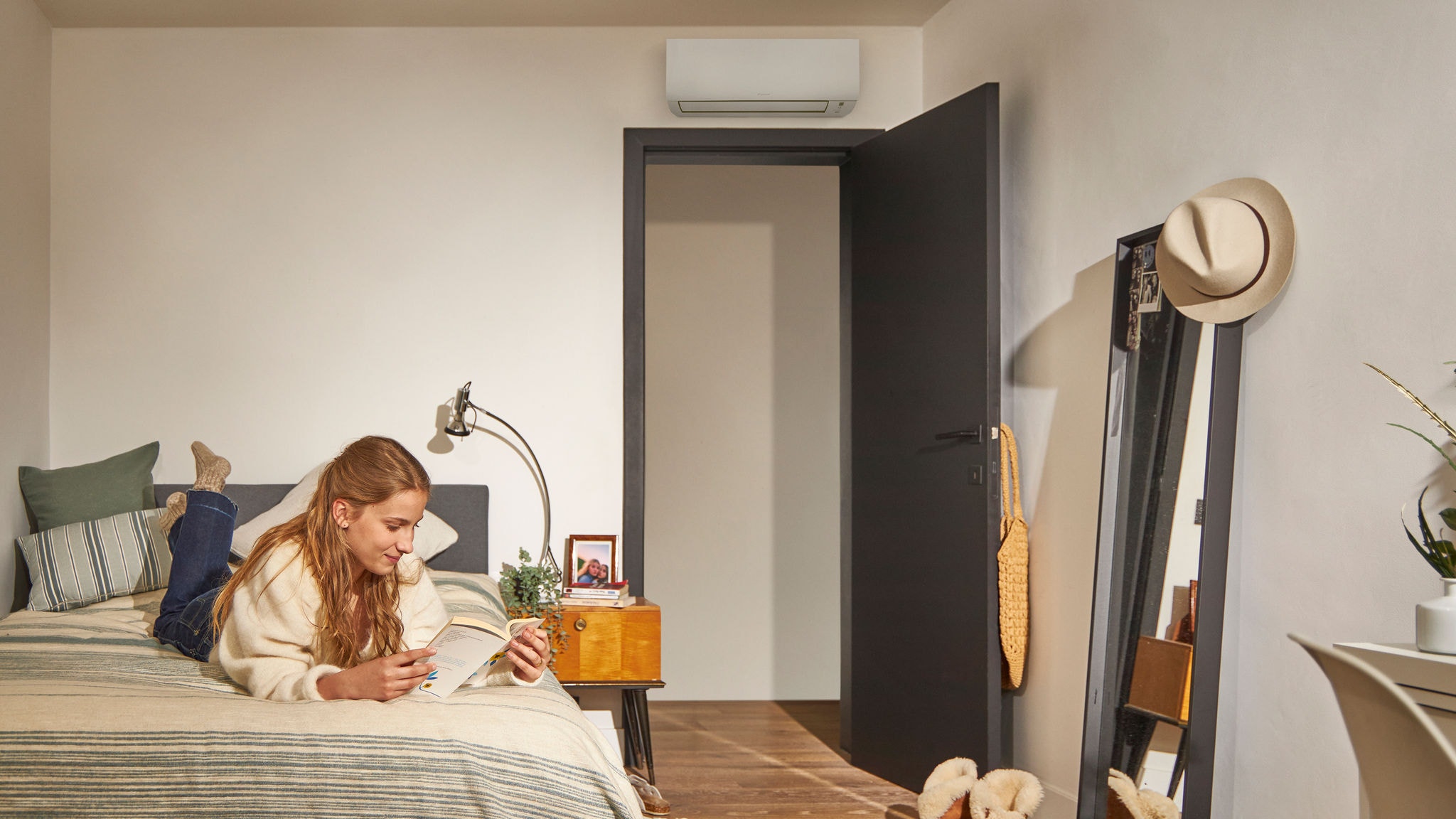 Mujer leyendo en la cama de un acogedor dormitorio con un equipo de aerotermia montado sobre la puerta, que garantiza una climatización energéticamente eficiente