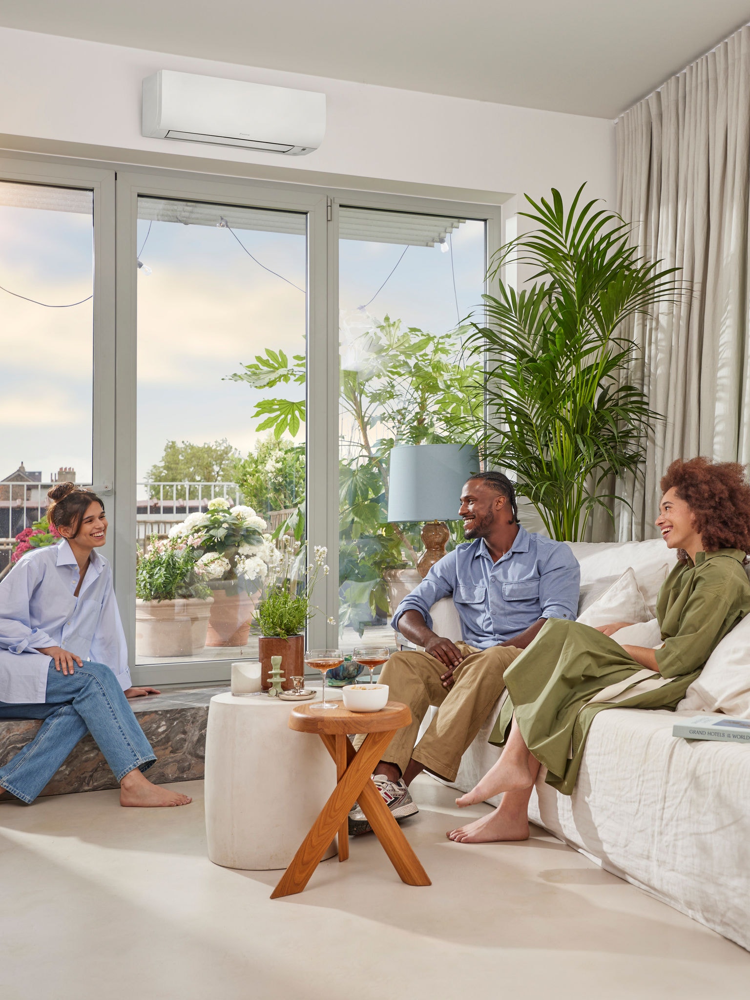 Un hombre y dos mujeres sentados y charlando en una estancia luminosa y espaciosa, con paredes blancas y un discreto equipo de aerotermia blanco montado en la pared, sobre un gran ventanal, en un día soleado