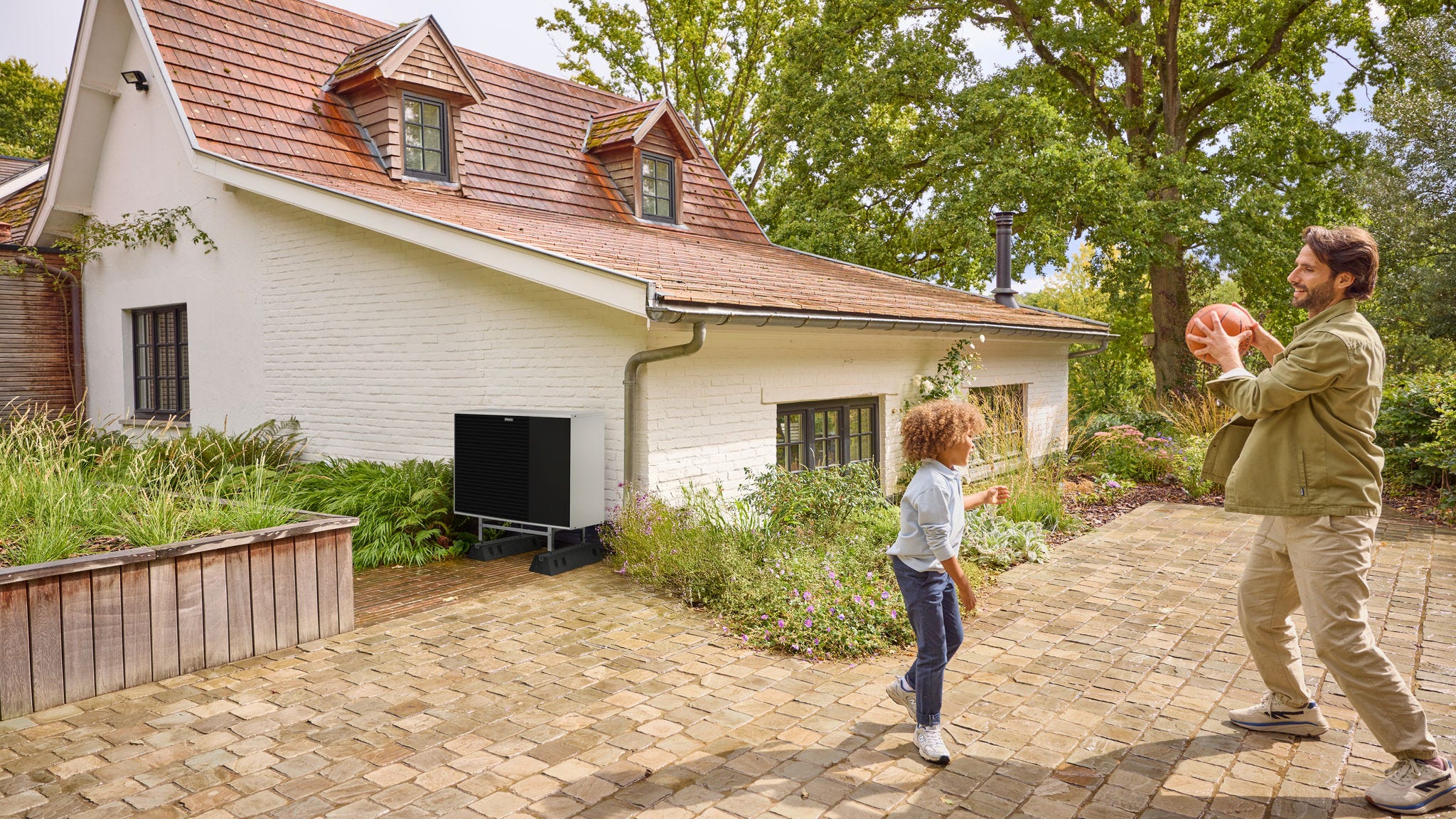 Padre jugando a la pelota con su hijo en el jardín, con un equipo de aerotermia instalado cerca