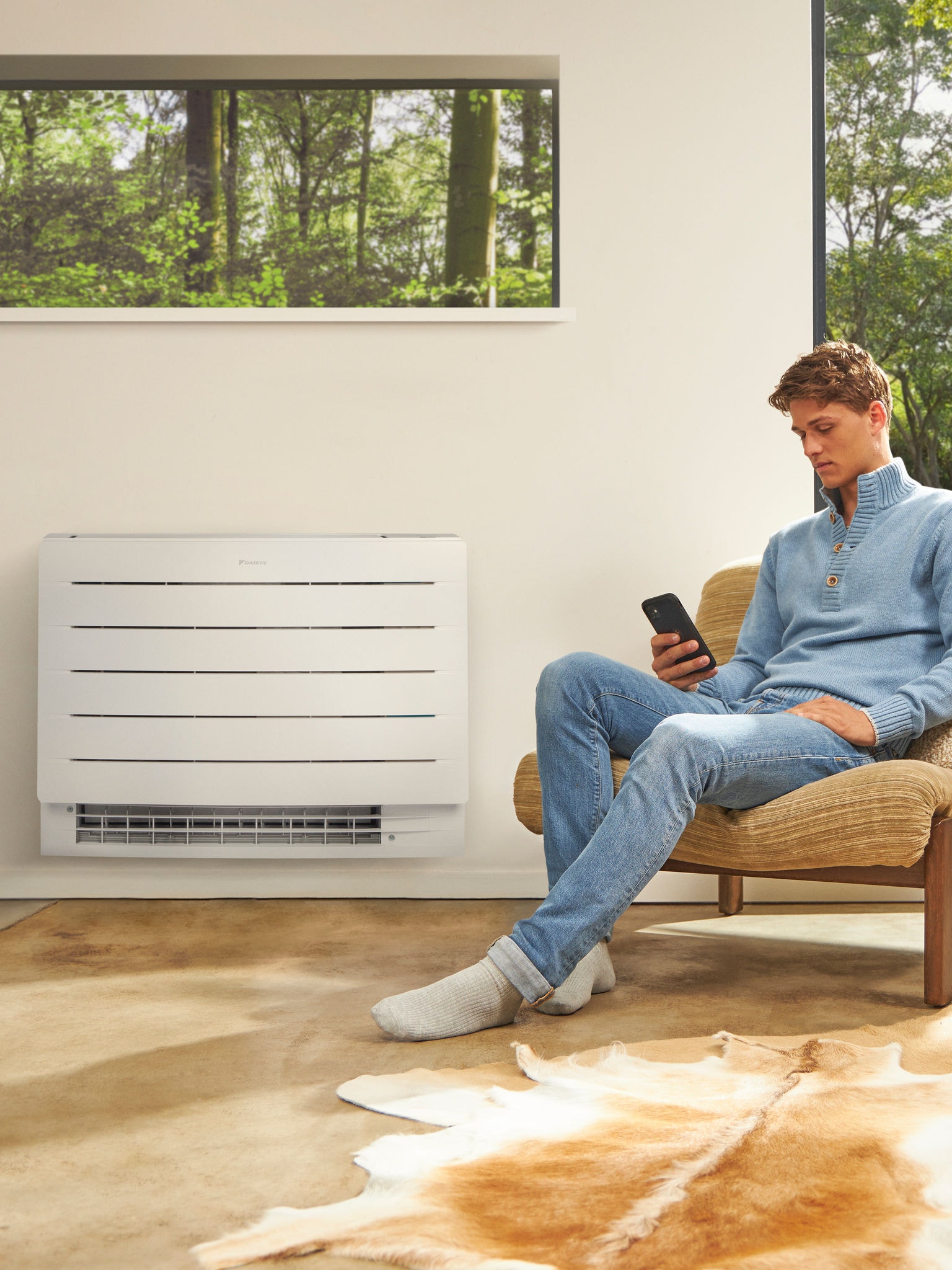 Un hombre sentado en una habitación luminosa mirando su teléfono con un equipo de aerotermia en la pared detrás de él