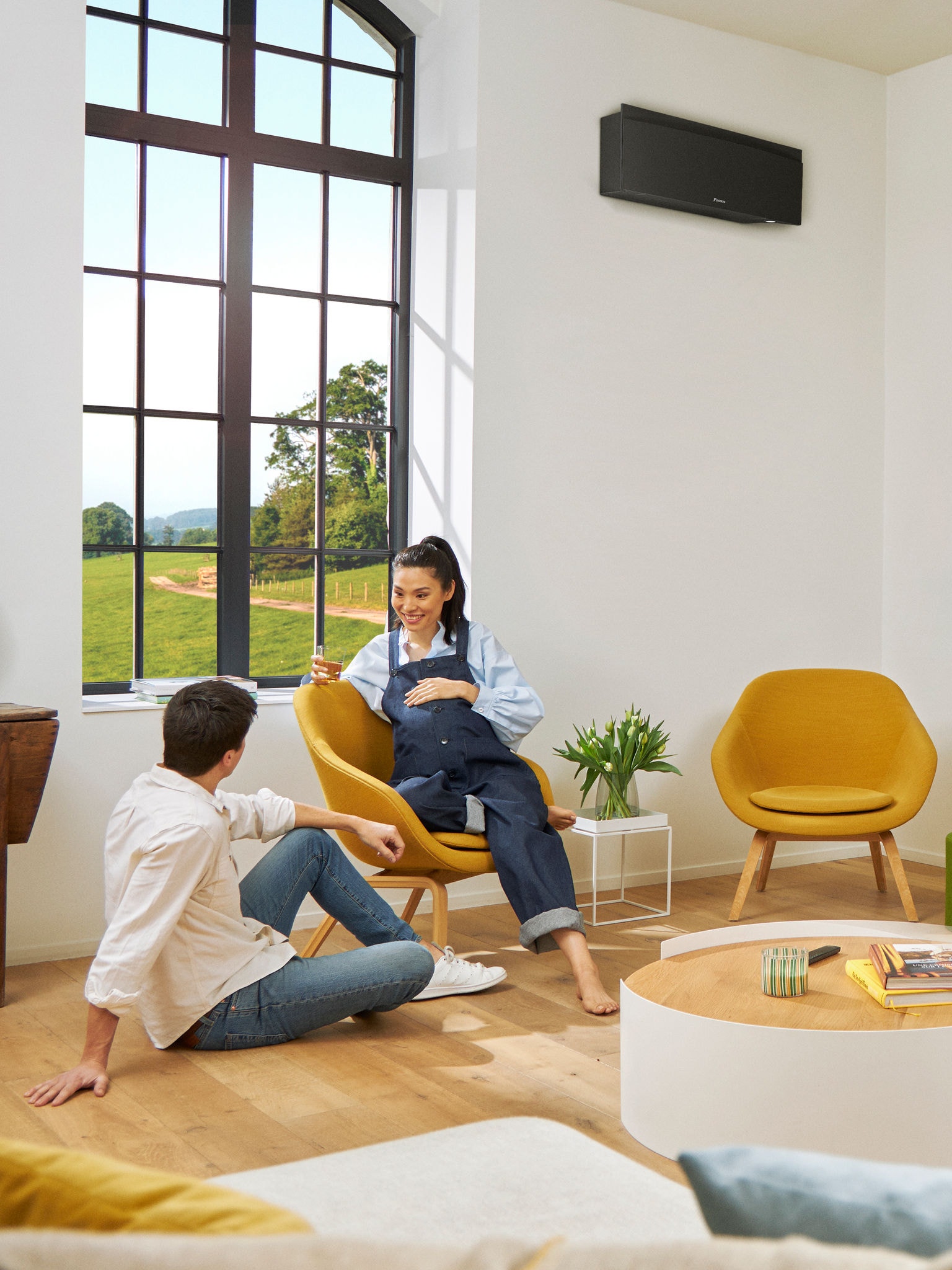 Un hombre y una mujer sentados frente a una gran ventana en una habitación luminosa con un equipo de aerotermia negro montado en la pared detrás de ellos