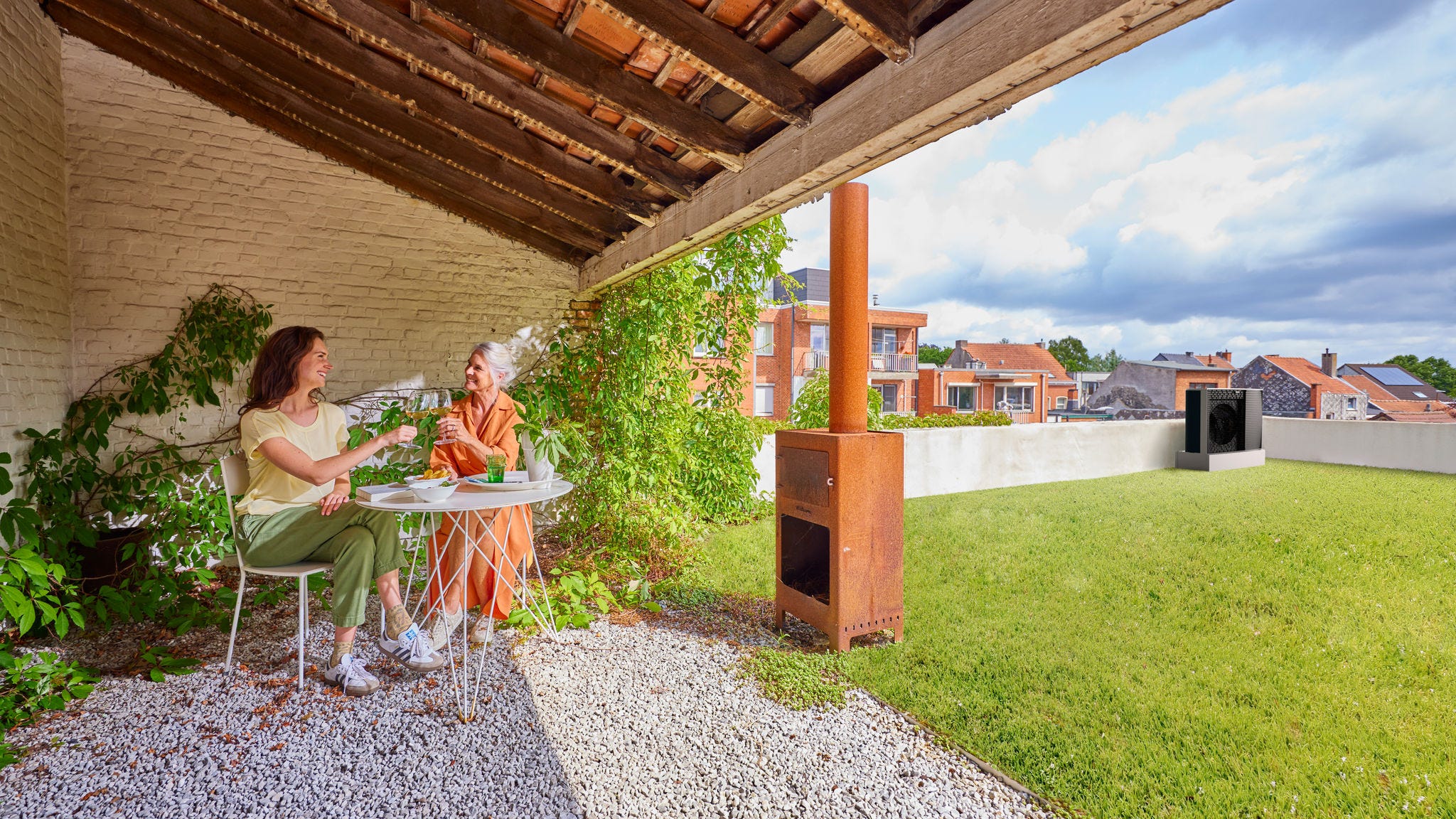 Una madre y una hija sentadas en una mesa al aire libre, bajo un techo inclinado de madera, junto a una estufa de leña
