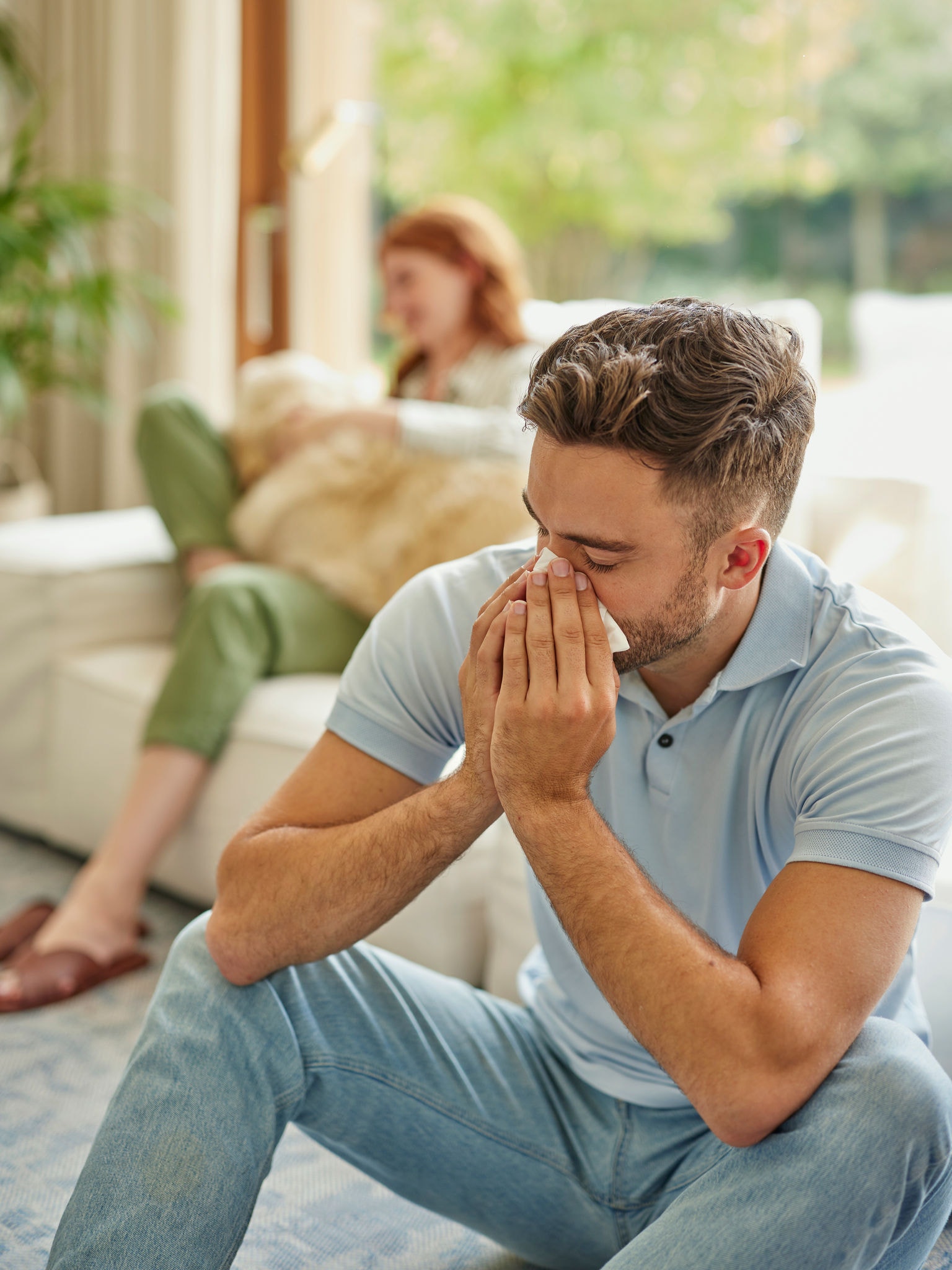 Mujer al fondo sentada en un sofá con un golden retriever en el regazo, hombre en primer plano estornudando
