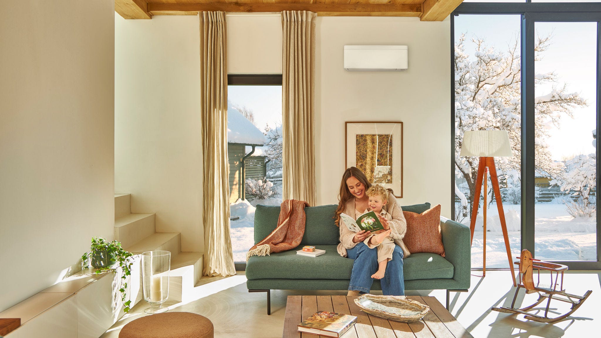 Mother sitting on the couch under a wall mounted air-to-air heat pump reading a children's book with her toddler on her lap enjoying the warmth while it's snowing outside